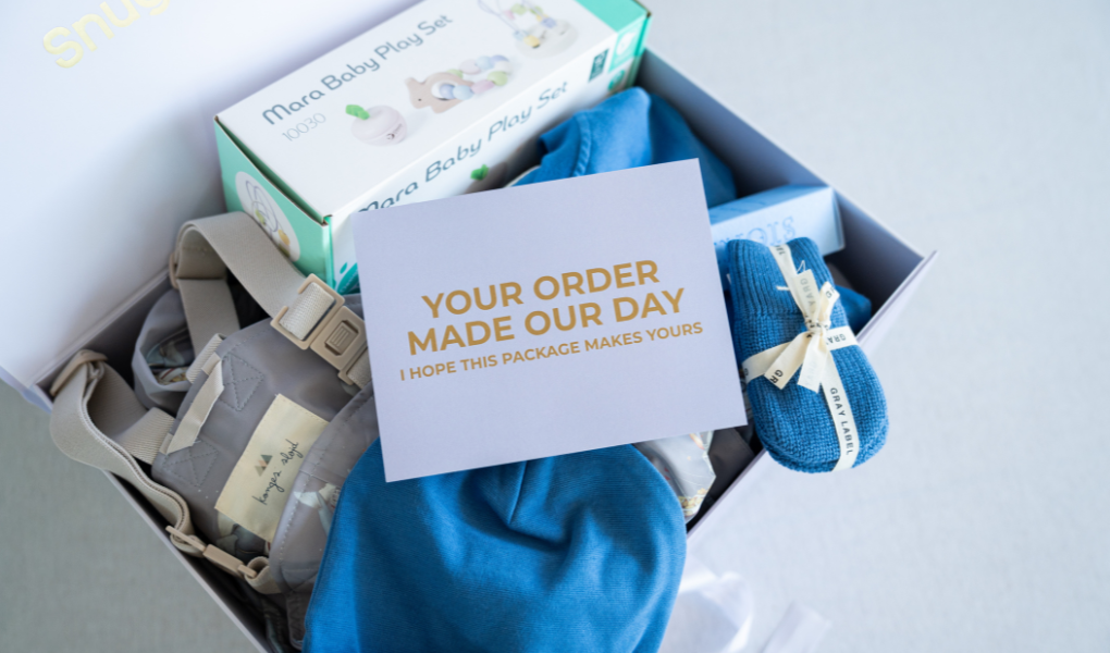 Baby gift set with blue socks, gray romper, and white card in a box.