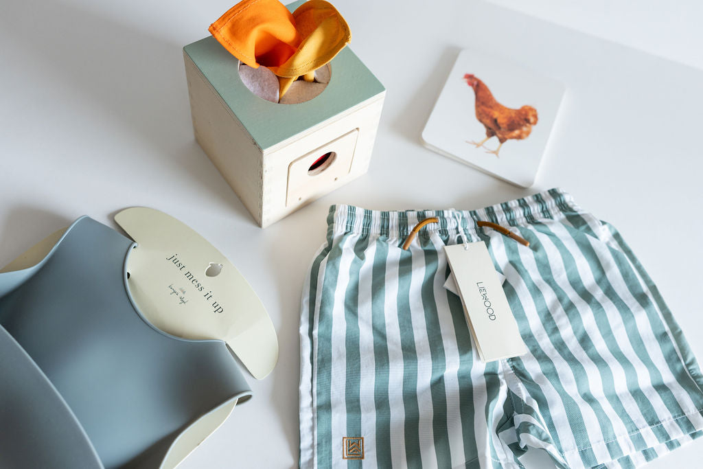Flat lay of high-quality baby boy products including a blue silicone bib, a wooden toy box with orange fabric, striped green and white Liewood swim shorts, and a chicken-themed learning card.